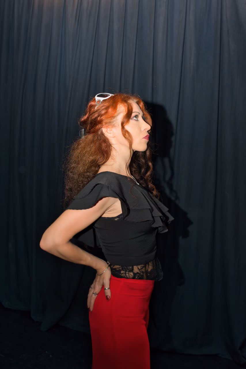 Eva ArtVision de perfil en escena, top de encaje negro y pantalón rojo, sobre fondo de telones oscuros